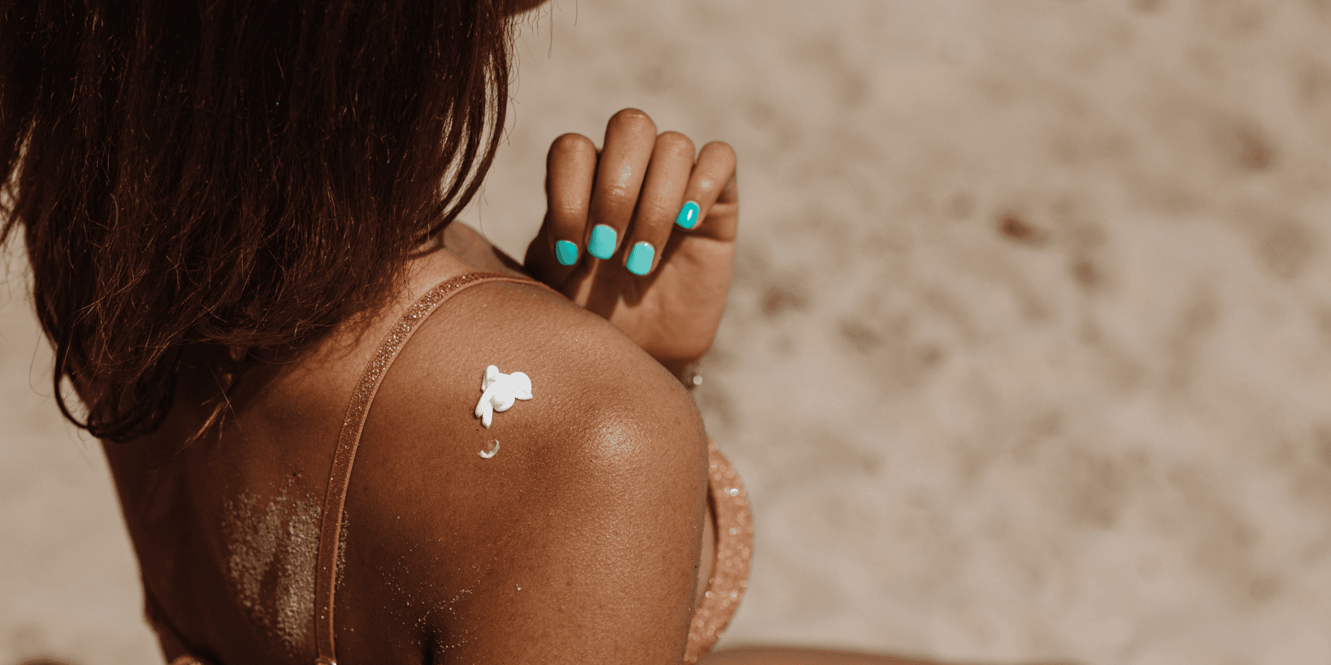 There is a woman with golden brown skin. She has her back to us and there is vegan sunscreen on her shoulder. Her hand is raised to her shoulder as if she is about to wipe the suncream in. Her nails are painted bright blue and you can see that she is on a beach because of the sand in the backgroun.