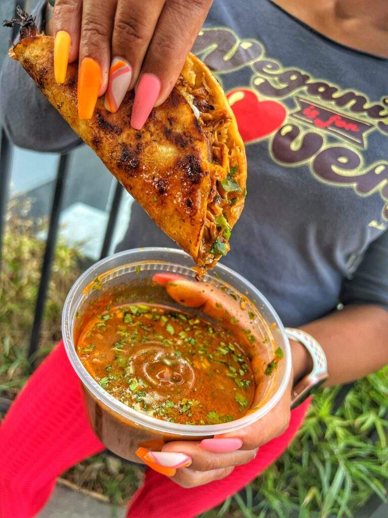 Someone dipping vegan quesabirria tacos into Mexican sauce at a vegan street food fair in the US
