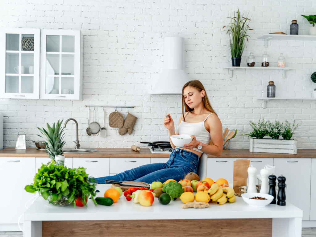 A woman eating vegan food for a healthier gut whilst sat in her kitchen with an array of plant-based foods on the kitchen counter
