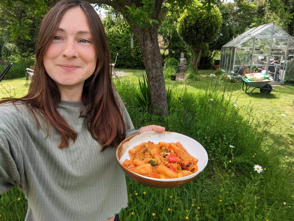 Alice is taking a selfie in a beautiful Suffolk garden whilst holding a bowl of allplants food in her other hand. She's smiling happily because the rigatoni smells so good!