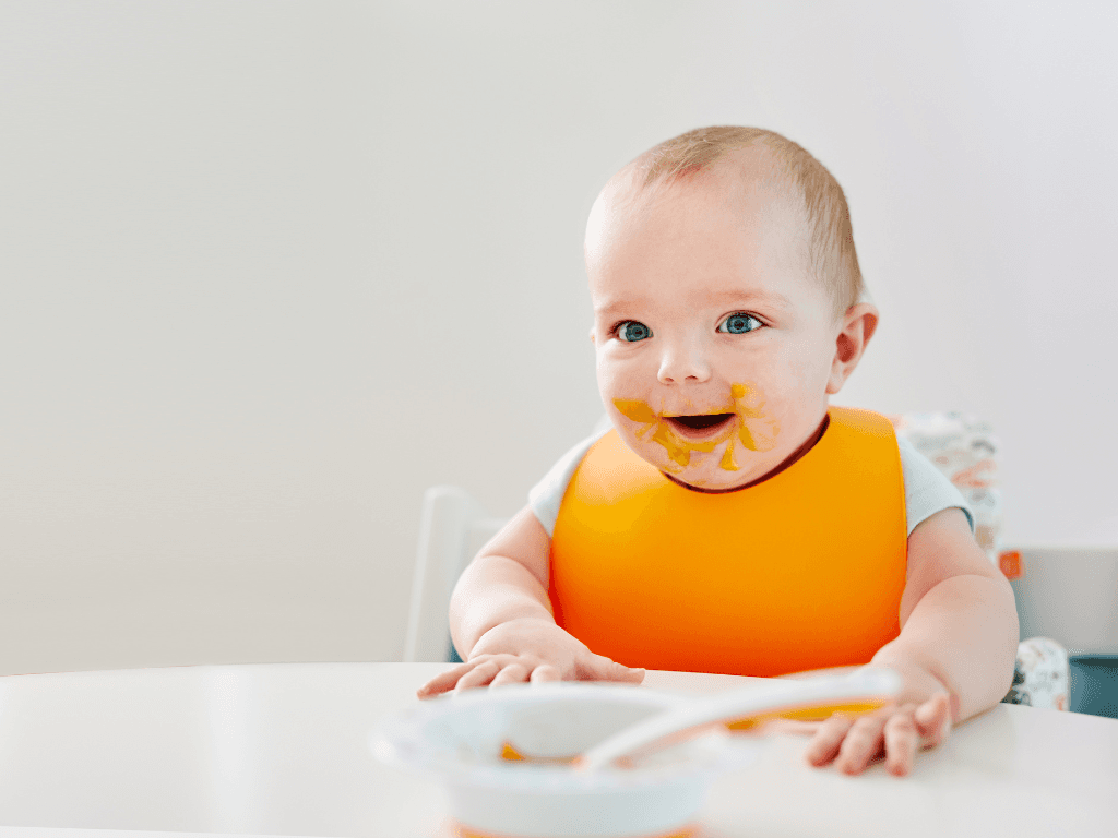 A baby wearing an orange bib sat at a table. The baby has vegan food all over his face.