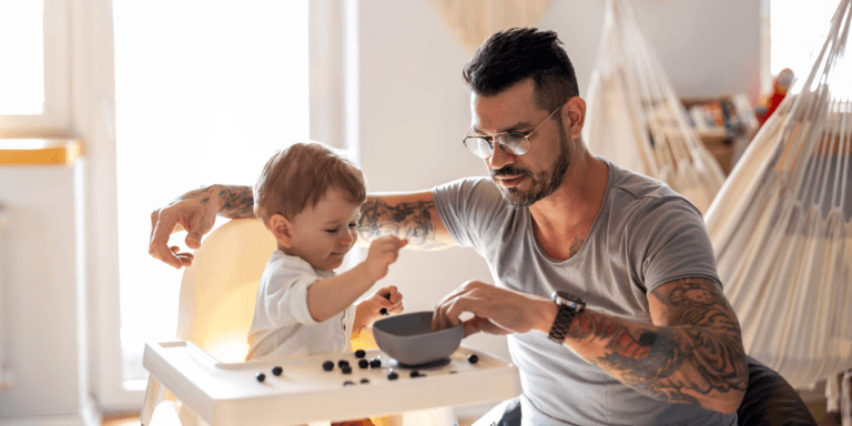 A father with tattoos on his arms feeding his vegan baby blueberries from a bowl