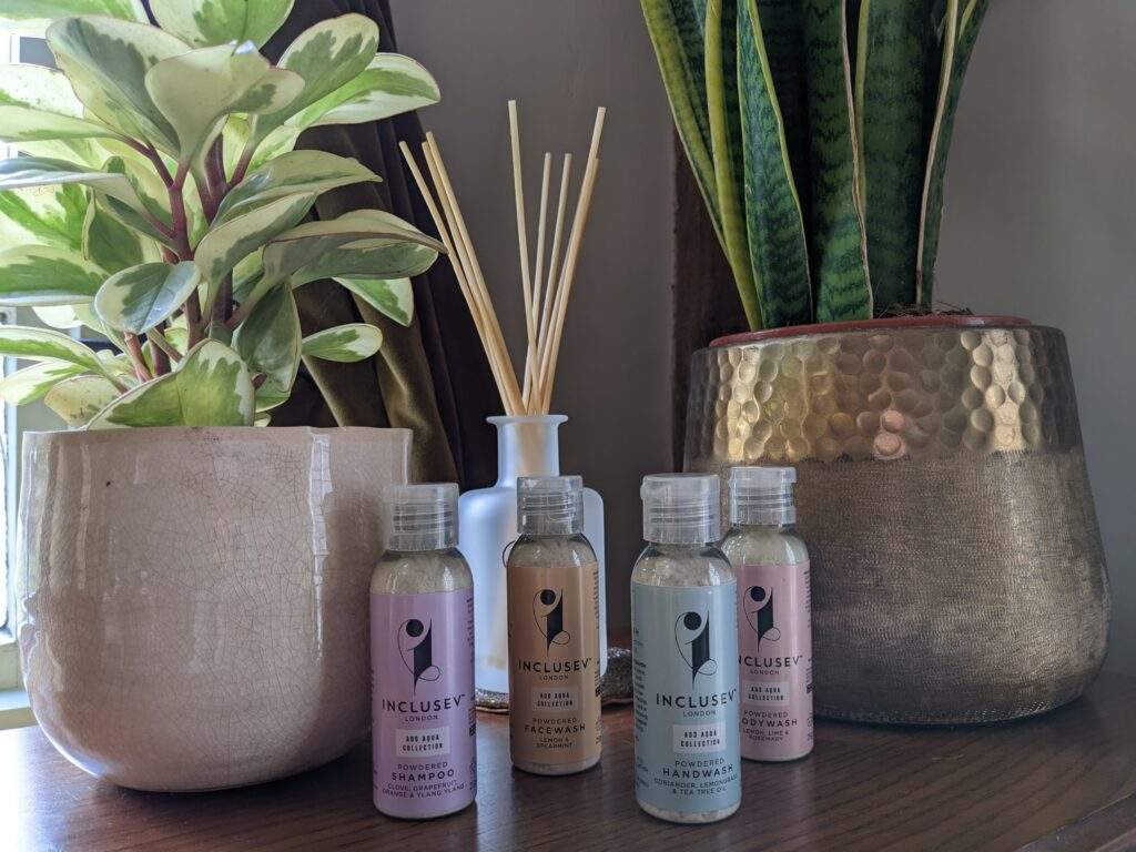 4 bottles of Inclusev's waterless beauty bottles. There are two plant pots with green leafy hosueplants in and a reed oil diffuser in the background