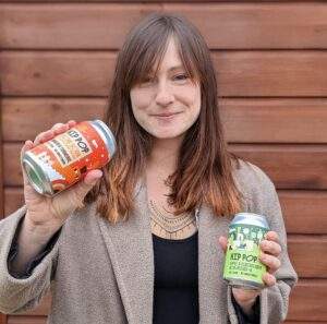 Alice is standing in front of a wooden background. She is wearing a camel coloured coat and a black vest. She is holding a can of kombucha in each hand. One is red and orange and the other is different shades of green. Alice has a smile on her face.