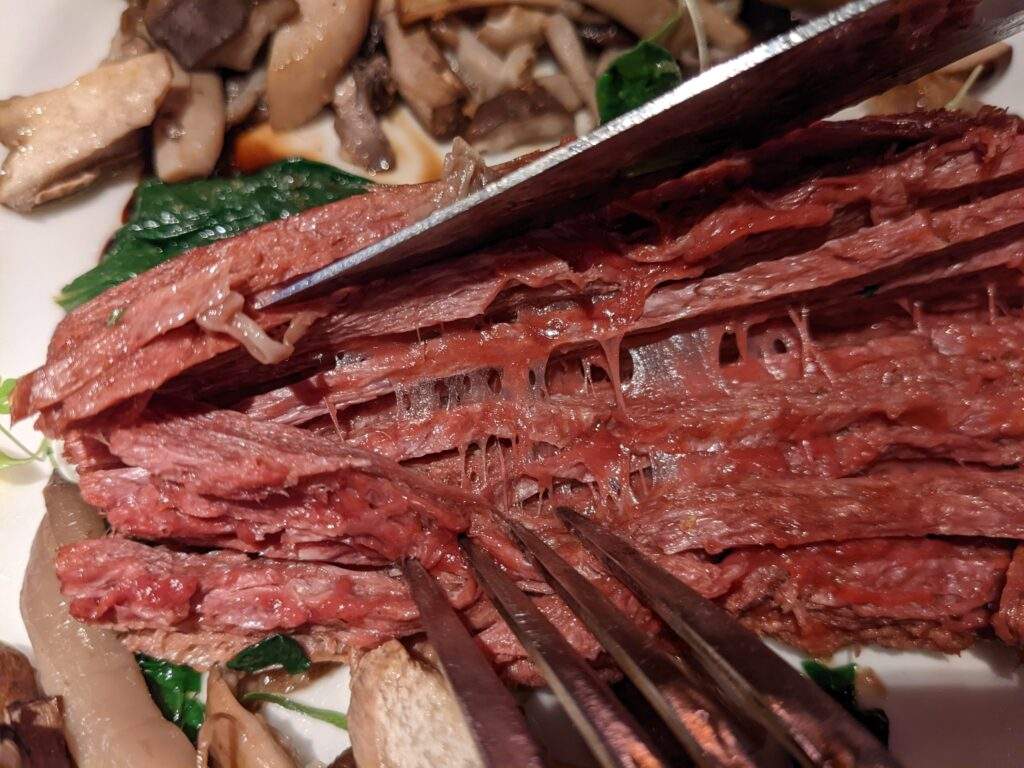 A knife and fork pulling apart the vegan fibres of 3D printed steak, revealing sticky sinews that seem to hold the fibres together.
