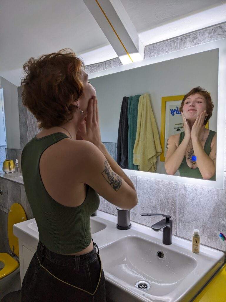 A woman massaging her face in circular motions. She is using a waterless beauty face wash to cleanse her pores. She has short auburn hair and has a tattoo on her arm.