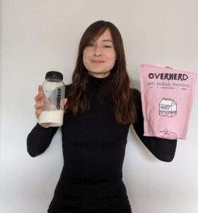 Alice is awkwardly holding a bottle of liquid oat milk in one hand and a pouch of Overherd powdered oat milk in the other. She's wearing a black turtle neck thermal top and her hair is long and brown. She's smiling.