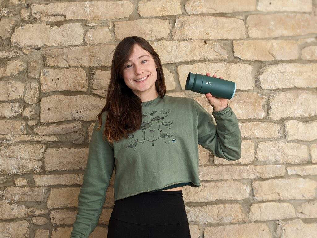 Alice and her eco-friendly thermos made out of stainless steel and recycled plastic saved from the ocean. She's stood in front of a brick wall and she has a big grin on her face.
