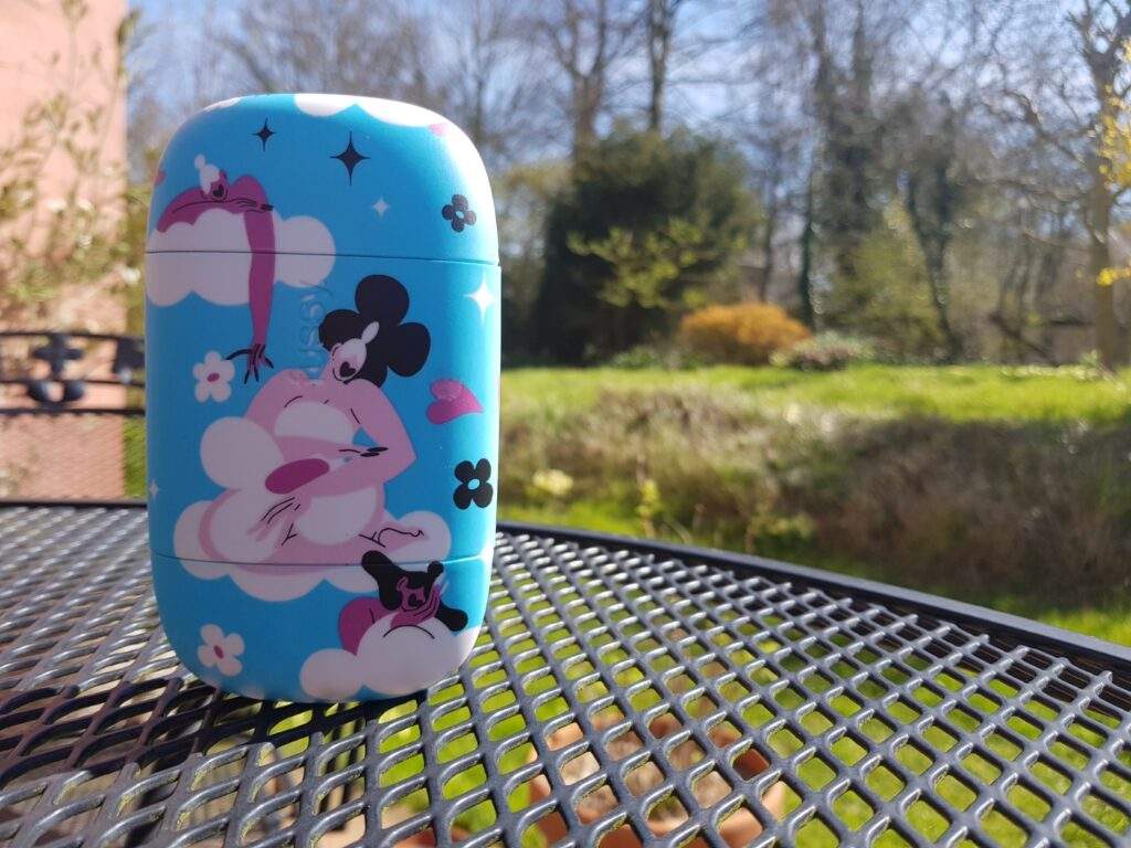Deodorising roll on inside a beautiful blue case with a dreamy cloud-filled design. It's stood up on a table in a garden. You can see trees and plants in the background.