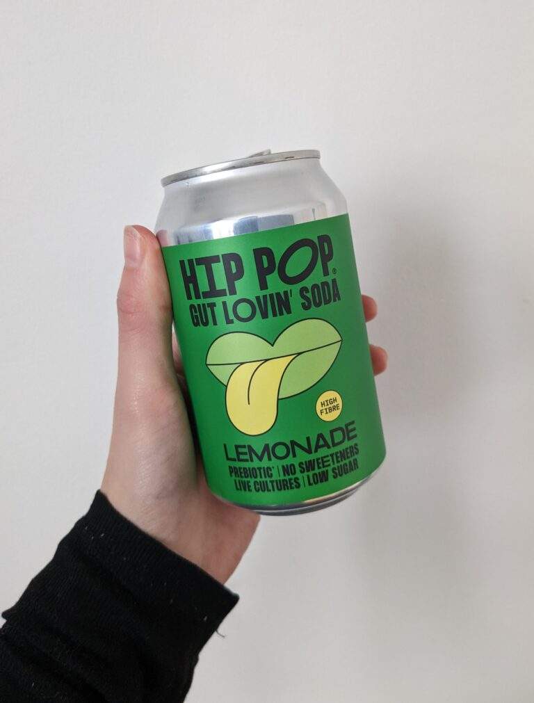 A hand holding a green aluminium can against a white background. It's a can of natural lemonade made by Hip Pop kombucha brand. THe branding shows a pair of licking lips.