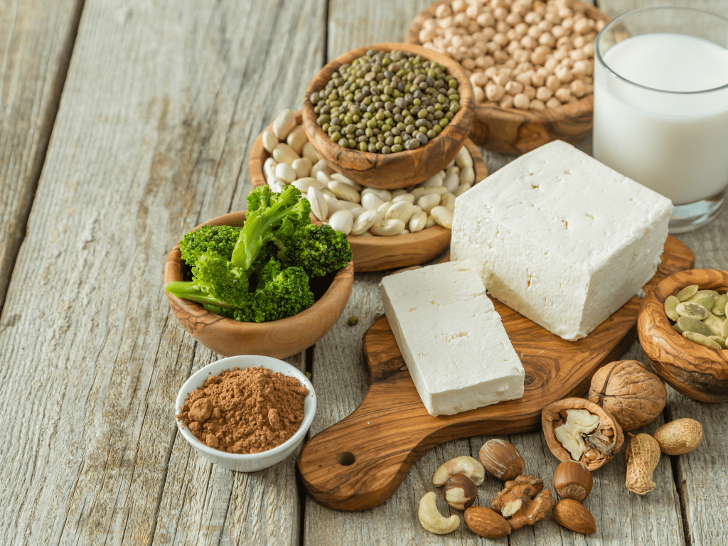 A selection of plant-based protein sources suitable for building muscle and weightlifting.. There are bowls of legumes (lentils and beans), broccoli, protein powder, and nuts. There's a wooden chopping board with a block of tofu on top and a glass of soy milk.