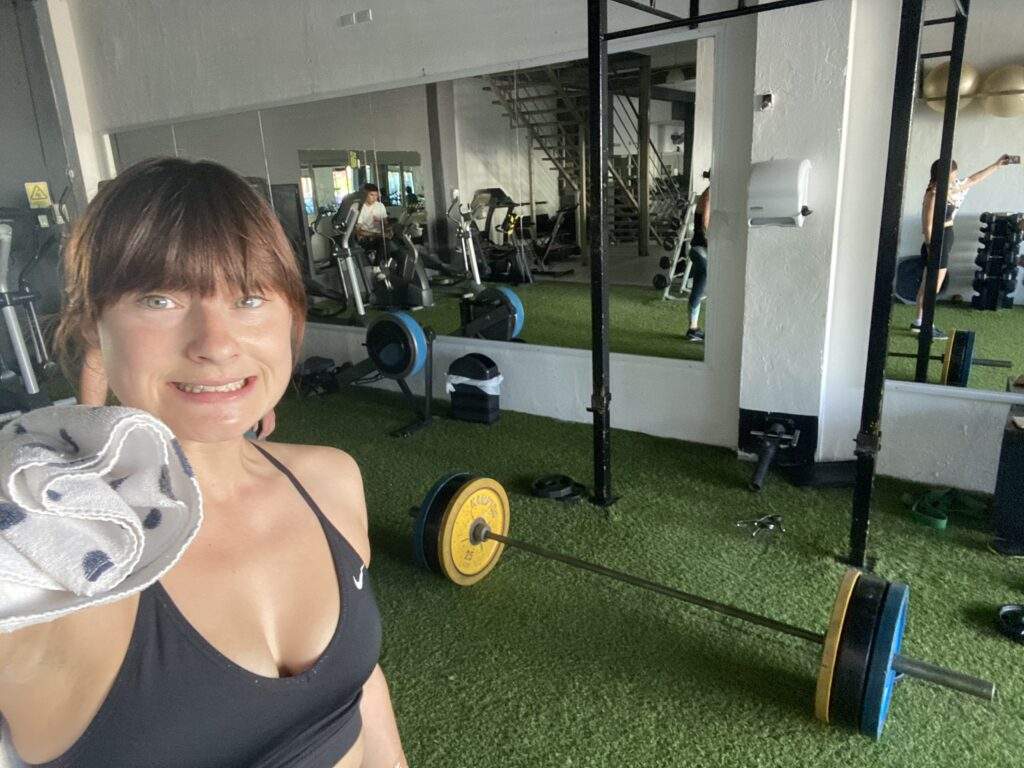 Sweaty vegan Alice taking a selfie in a Mexican gym. There's a deadlift barbell setup in the background.