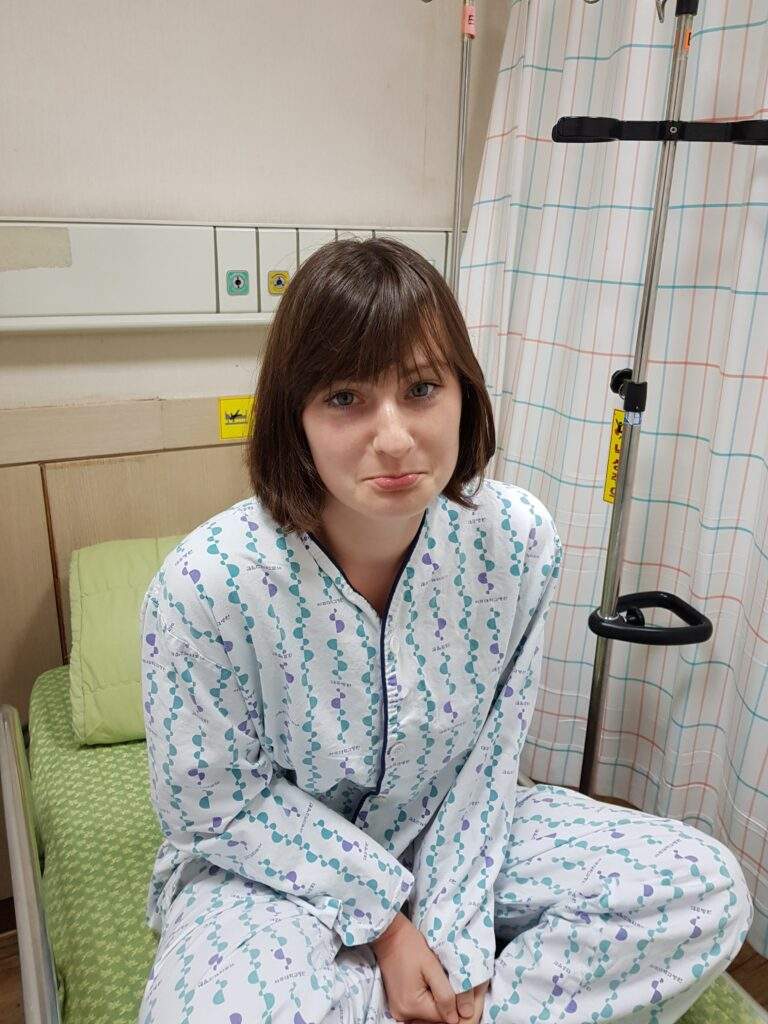 Alice is sat cross-legged on a hospital bed in A&E. She's wearing hospital PJs and is sticking her bottom lip out