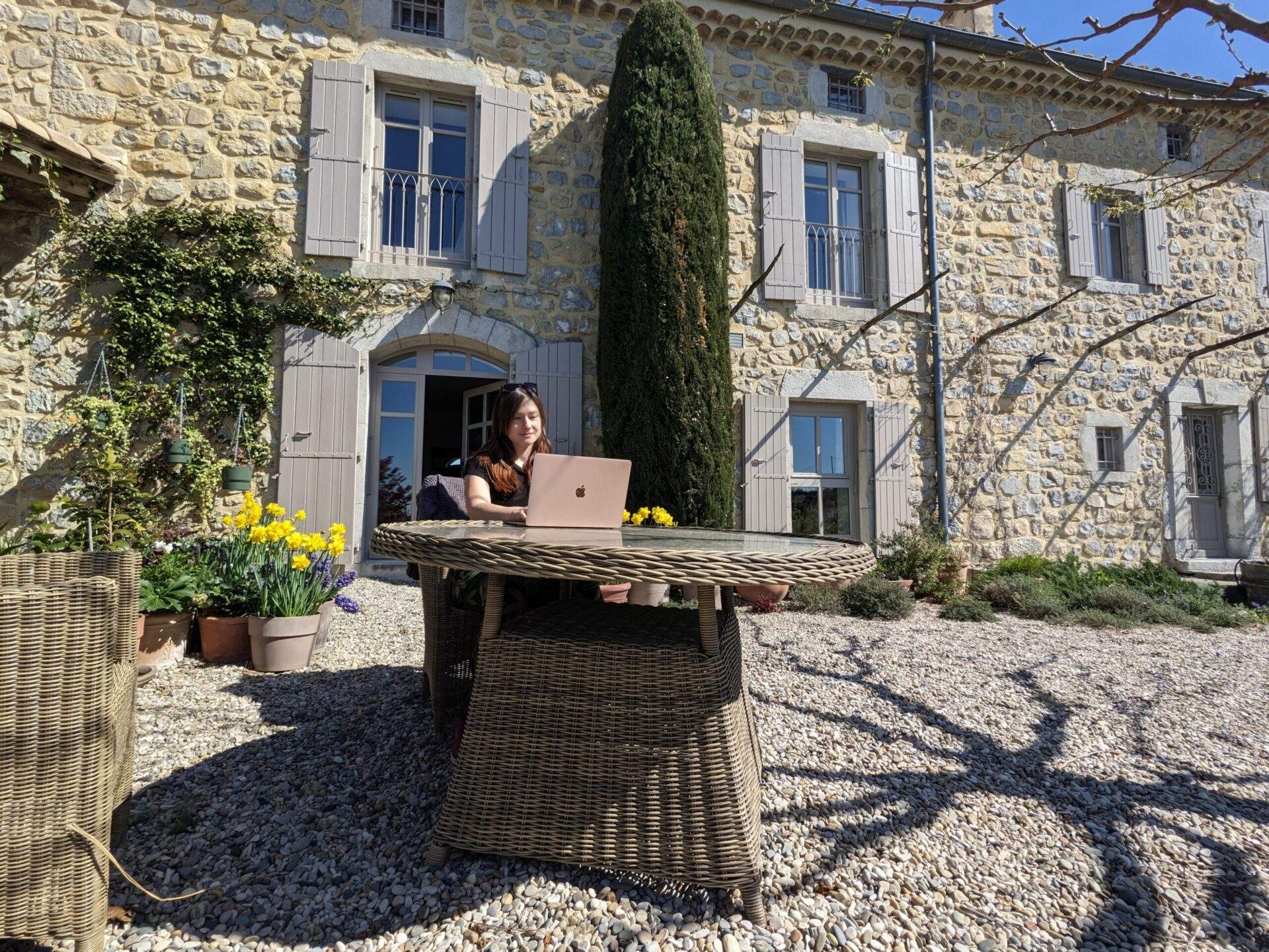 Alice is sat at a wicker table with her Apple Macbook Air. She's working in the sunshine in front of a house she is sitting in the Ardeche in France