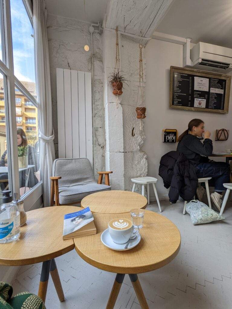 A mug of oat milk cappuccino is on a saucer on a table, next to a Jane Austen book. The setting is a cafe called Brumes, which serves speciality coffee. The decor is whitewashed with plants. You can just see blue sky out of the window on the left and there are women sat inside and outside of the cafe.