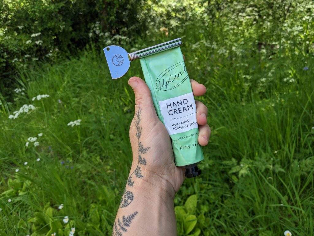 A hand holding a tube of hand cream by UpCircle Beauty. The tube has a metal twister on the top to help squeeze out every last drop of cream. In the background you can see wild flowers and long grass.