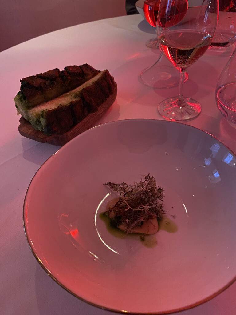 A big bowl with a blob of Aïgo Boulido on top of wild garlic infused oil and crispy moss balanced on top. There is a plate of wild garlic focaccia next to it and you can see the bottom of a wine glass. The Gauthier Soho restaurant is lit with red ambient lighting