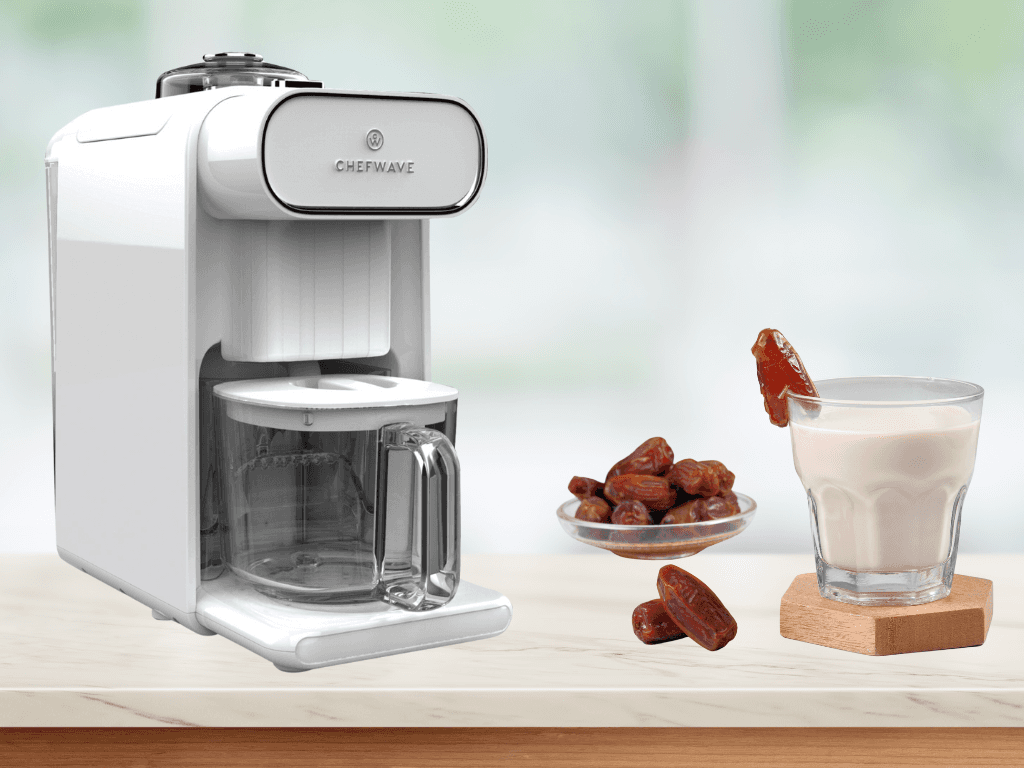 The ChefWave nut milk maker machine on a kitchen worktop. There's a glass of nut milk on the counter with a bowl of dates and some more dates scattered on the table.