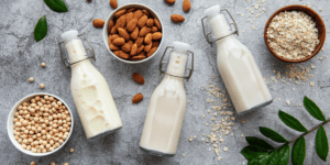A selection of homemade plant milks in glass bottles. They're laid out on a grey surface and surrounded by bowls of soybeans, almonds and oats. There are a few other plant-based ingredients scattered around as well.