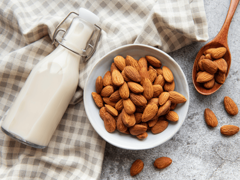 A bottle of almond milk on a tea towel with a bowl of almonds and a wooden spoon of almonds