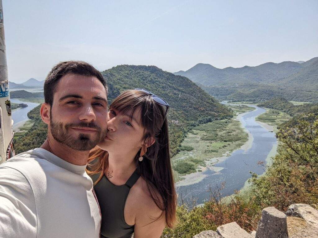 Alice and her partner Luka are standing at Pavlova Strana Viewpoint, overlooking the Green Pyramid hill and the bend in Rijeka Crnojevica. The river is beautifully blue and the shores are luscious green
