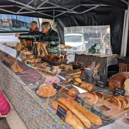 Two women stood behind a bakery stall in old town market in Annecy. There is an array of pastries and breads underneath a glass counter. A woman with a pink coat is stood in front of the stall being served
