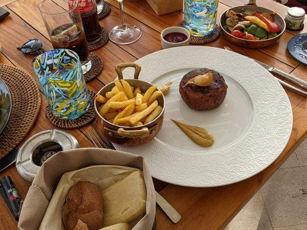 A Beyond Meat vegan steak with a mustard dressing and fries. There's a bread basket and grilled vegetables alongside and a few glasses of drinks (wine, coca cola, water, etc.) on the table.