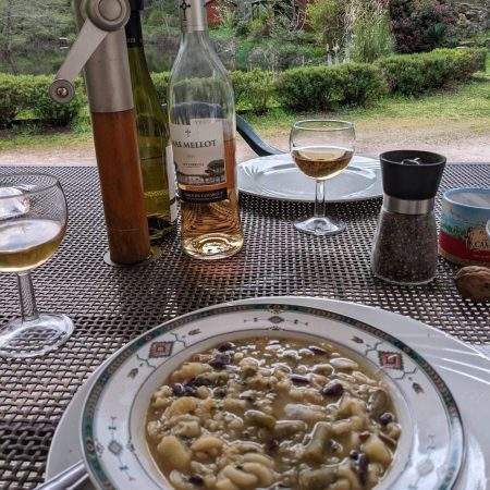 A bowl of Pistou soup, a Provencal dish that's traditionally vegan. The bowl is on a laid table with wine glasses and a bottle of natural vegan wine. There's a pepper grinder and in the background, you can see the bushes and trees that make up the gites of the moulin in Beynat